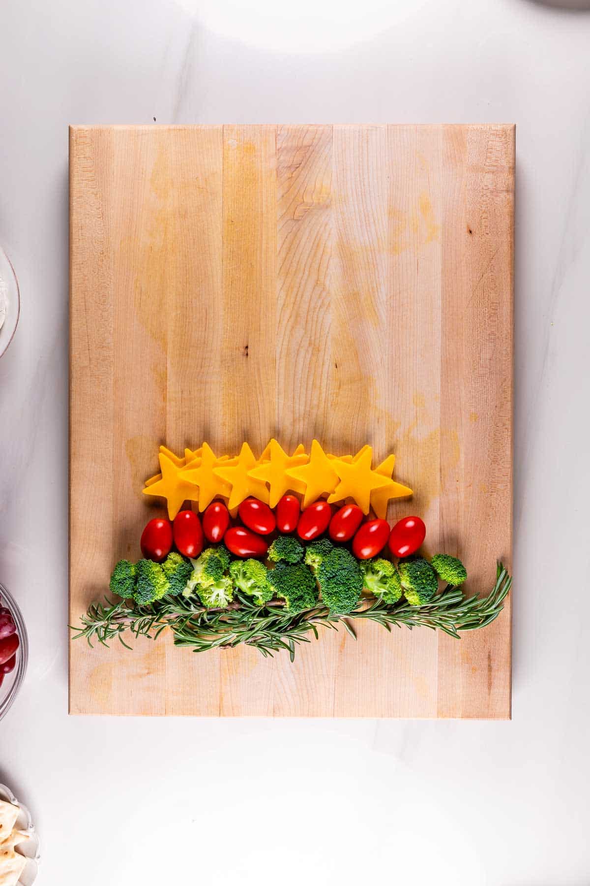 A wooden cutting board with sliced cheddar cheese stars, cherry tomatoes, broccoli florets, and sprigs of rosemary, arranged horizontally.