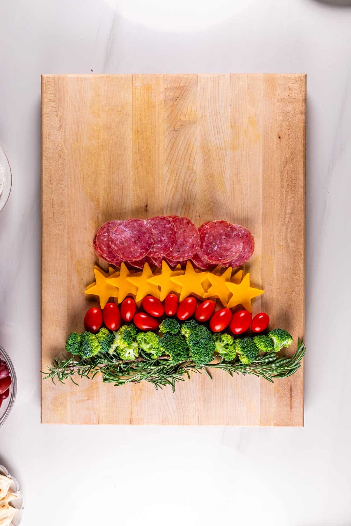 A cutting board with layered rows of salami, cheese cubes, cherry tomatoes, broccoli florets, and sprigs of rosemary, arranged in a colorful, organized pattern.