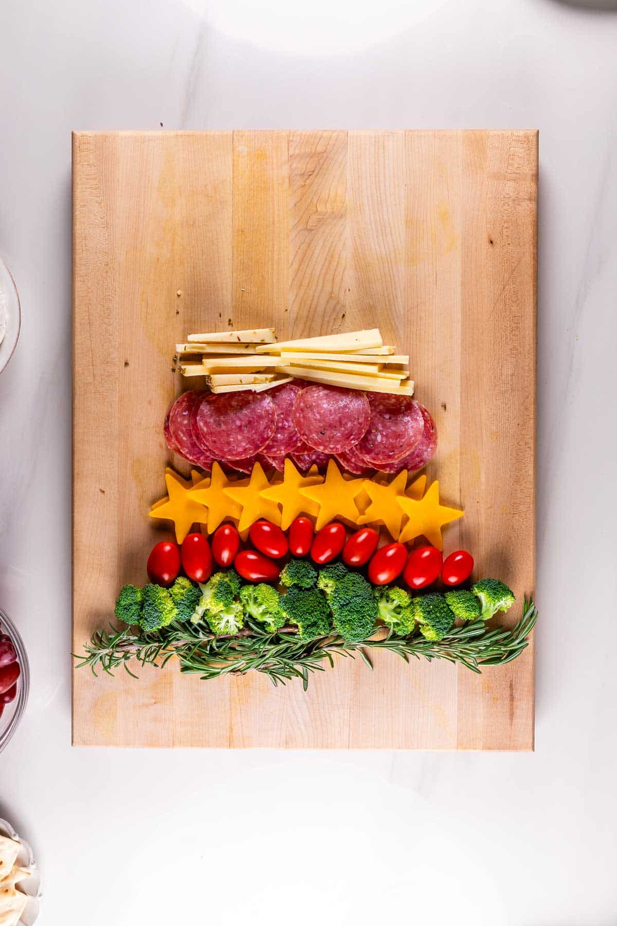 A wooden board with food arranged in the shape of a Christmas tree: breadsticks at the top, followed by a layer of salami, star-shaped cheese slices, cherry tomatoes, and broccoli, with sprigs of rosemary at the base.