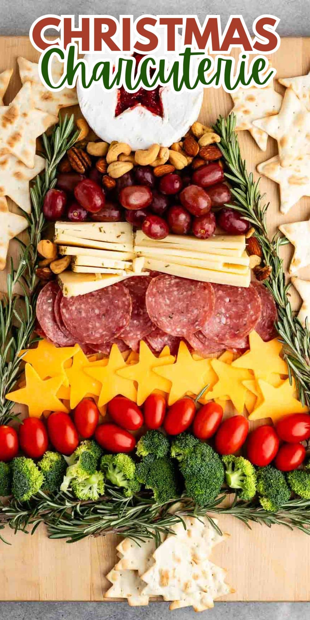 A festive Christmas charcuterie board arranged in the shape of a tree, featuring cheeses, meats, grapes, tomatoes, broccoli, and rosemary sprigs, surrounded by star-shaped crackers on a wooden board.