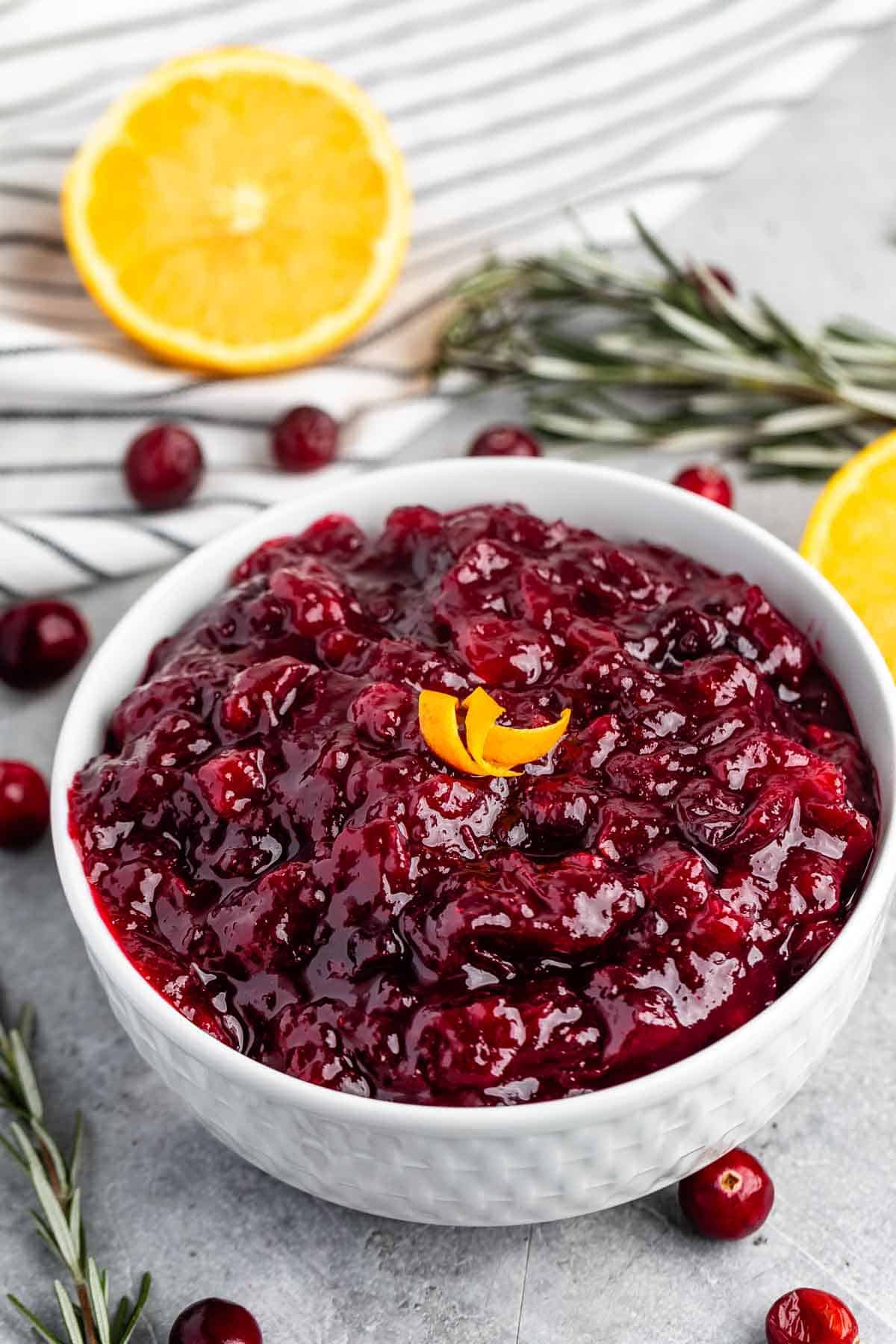 A white bowl filled with cranberry sauce garnished with orange peel sits on a table. Fresh cranberries, halved oranges, rosemary sprigs, and a striped cloth surround the dish.