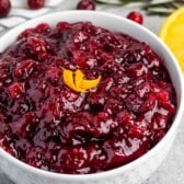 A white bowl filled with glossy cranberry sauce garnished with orange zest curls. Fresh cranberries and orange slices are scattered around on the gray surface. Sprigs of rosemary are nearby, adding a touch of green.