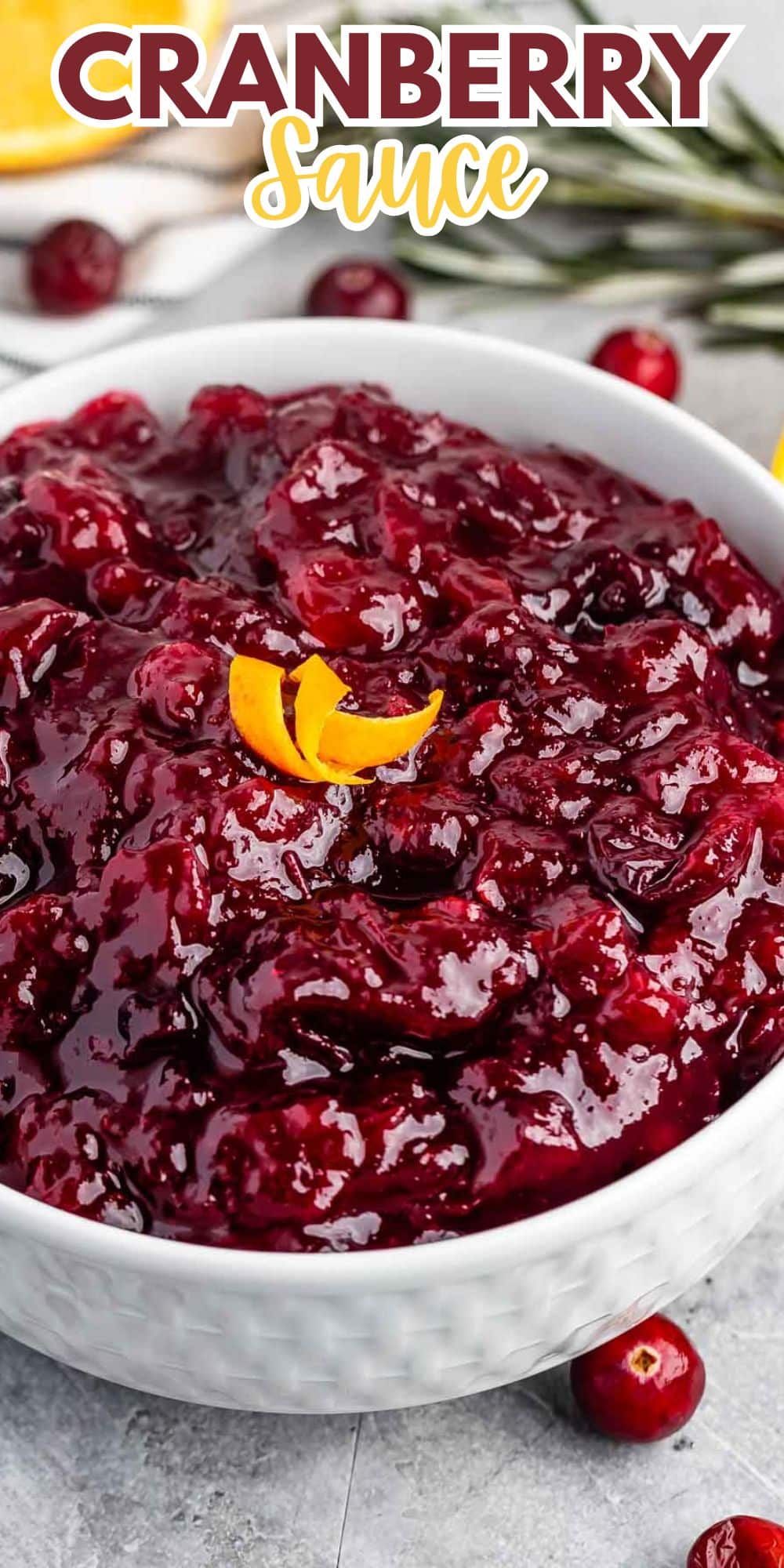 A bowl of vibrant cranberry sauce, topped with a small twist of orange peel. In the background, whole cranberries and a sprig of greenery are visible. The words Cranberry Sauce are written at the top in bold text.