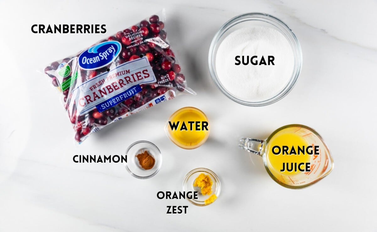 A top view of cranberry sauce ingredients: a bag of cranberries, a bowl of sugar, a glass of water, a cup of orange juice, a small bowl of cinnamon, and a dish of orange zest, all labeled on a white surface.