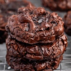 A close-up of a stack of three decadent chocolate cookies on a cooling rack. The cookies are rich and glossy, with a slightly cracked surface, evoking a freshly baked, indulgent treat.