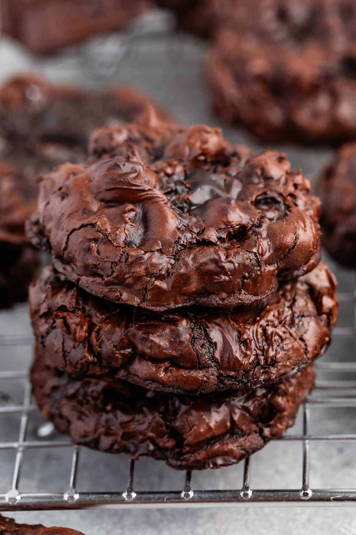 A close-up of a stack of three decadent chocolate cookies on a cooling rack. The cookies are rich and glossy, with a slightly cracked surface, evoking a freshly baked, indulgent treat.