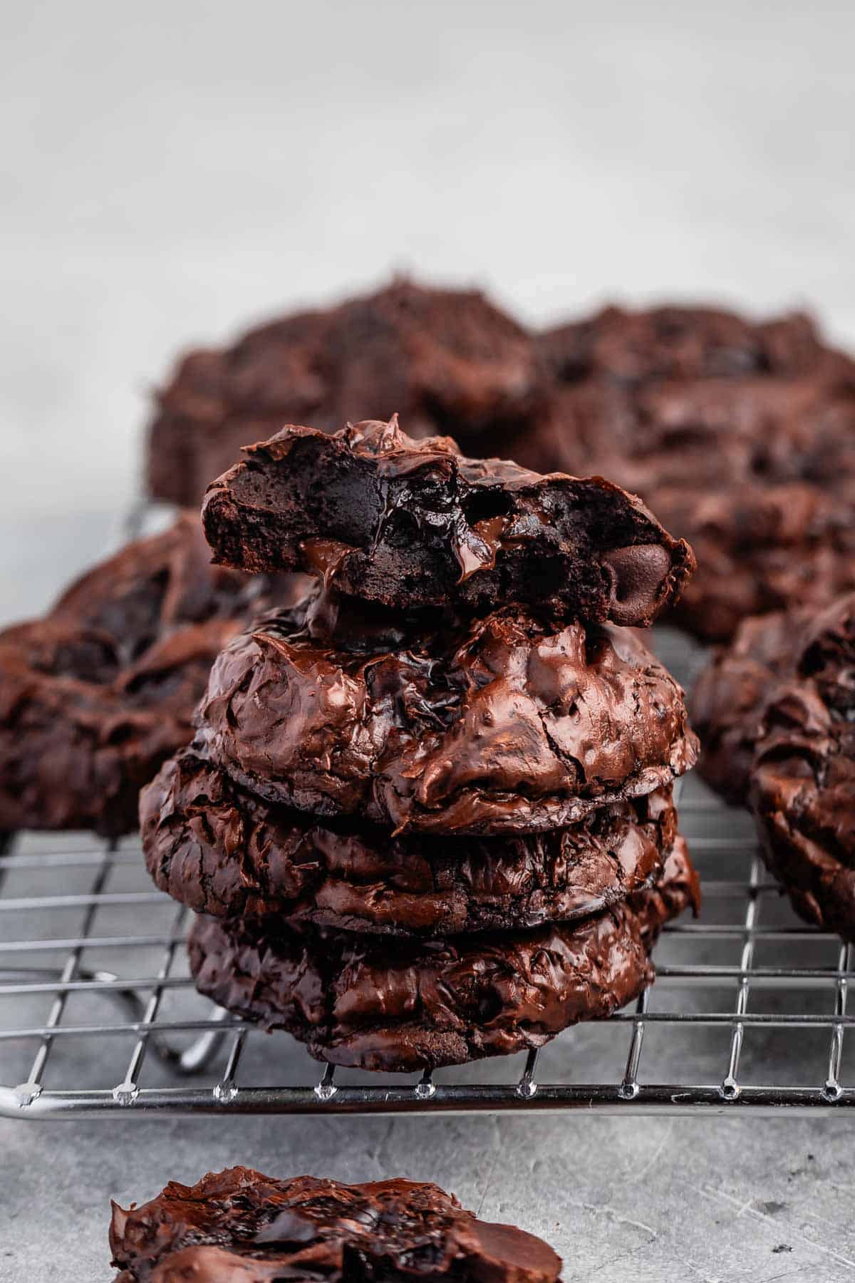 A stack of three rich, chocolate brownie cookies sits on a cooling rack. The top cookie is broken in half, showcasing a gooey, fudgy interior. More cookies are scattered in the background on a light gray surface.