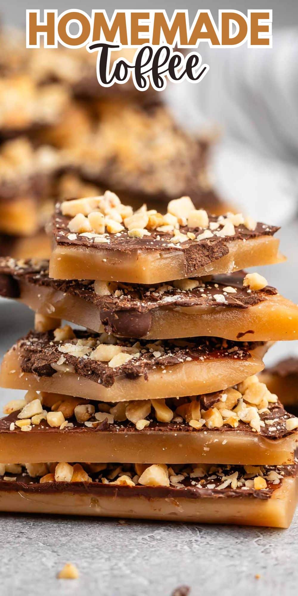 A stack of homemade toffee pieces topped with chopped nuts on a gray surface. The toffee showcases layers of caramel and chocolate with a sprinkle of nuts. The text HOMEMADE toffee is visible at the top.