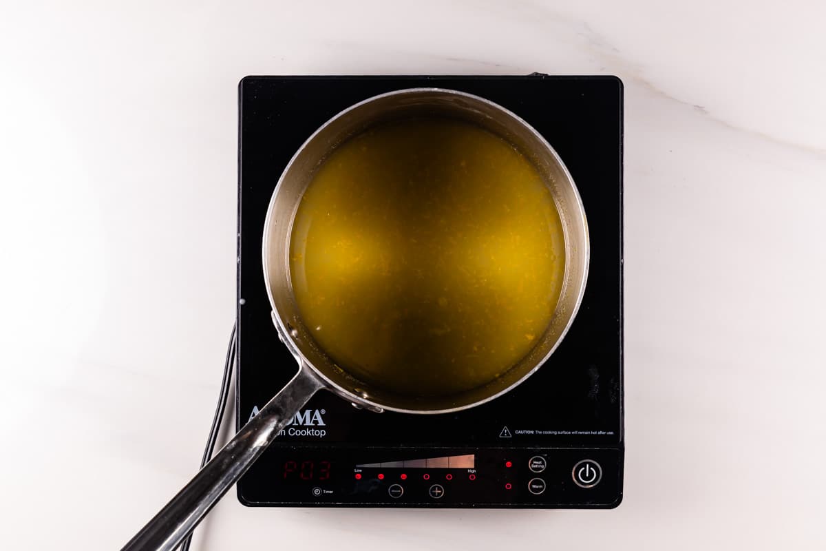 A pot of golden yellow liquid is on top of a black induction cooktop. The cooktop displays a digital reading. The scene is set against a smooth white countertop, and a metal ladle rests inside the pot.