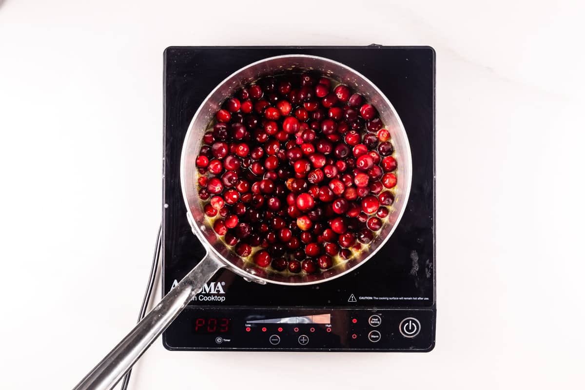 A pot of cranberries is being heated on an induction cooktop. The cooktop displays a temperature setting. The cranberries are submerged in liquid, indicating they are being cooked, possibly for a sauce or compote.