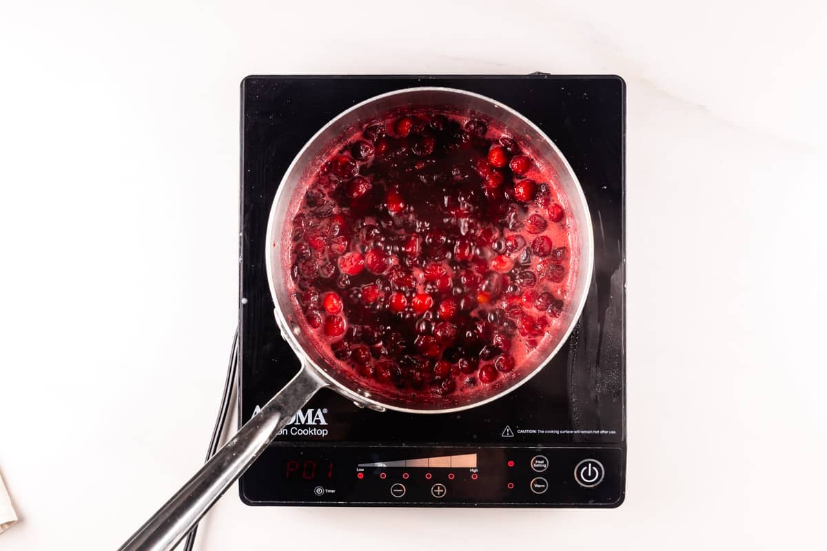 A saucepan filled with a bubbling red cranberry sauce placed on a black induction cooktop. The sauce contains visible whole cranberries and is in the process of simmering.