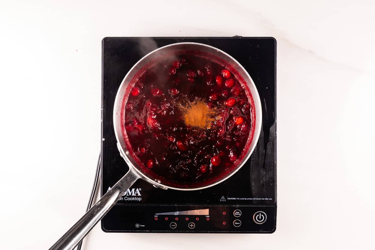 A pot of cranberry sauce with visible cranberries and a sprinkle of cinnamon on top, simmering on an induction stove. The surface is white, and the steam rises from the pot, indicating its hot.