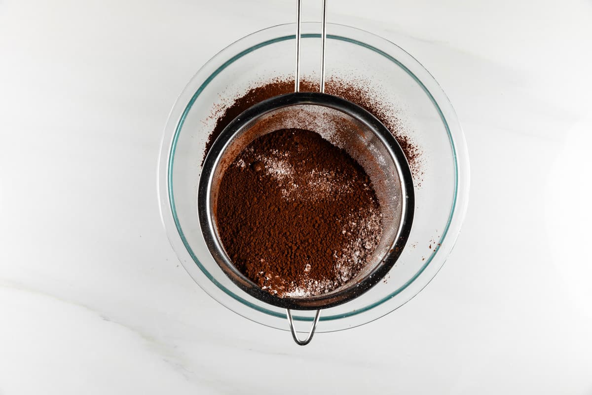 A glass bowl with a metal sieve containing sifted cocoa powder and flour, on a light marble surface.