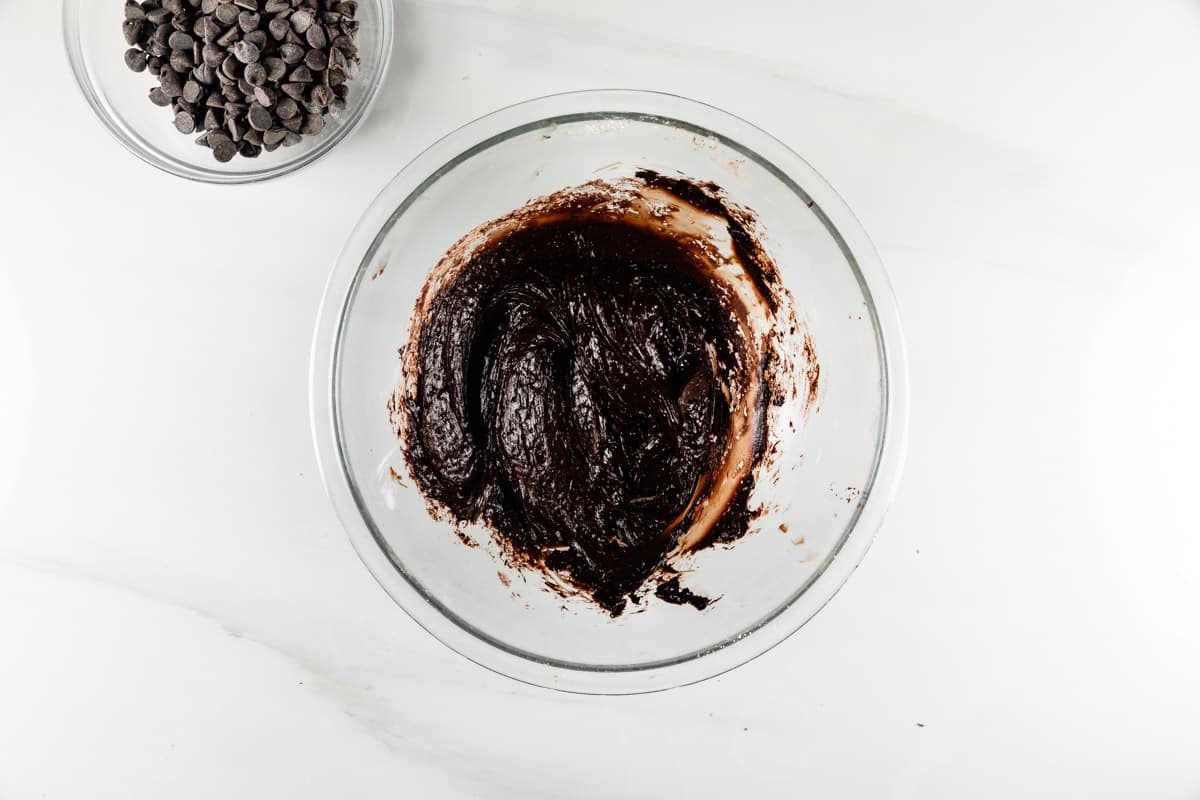 A glass bowl filled with rich, dark chocolate batter on a white marble surface. A small bowl of chocolate chips sits nearby, ready to be added.