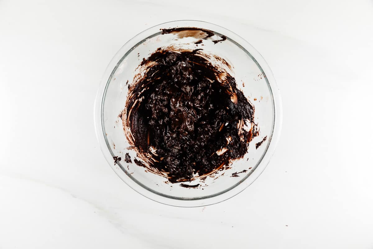 A glass bowl containing thick, dark chocolate brownie batter on a white surface. The batter has been stirred and clings to the sides of the bowl.