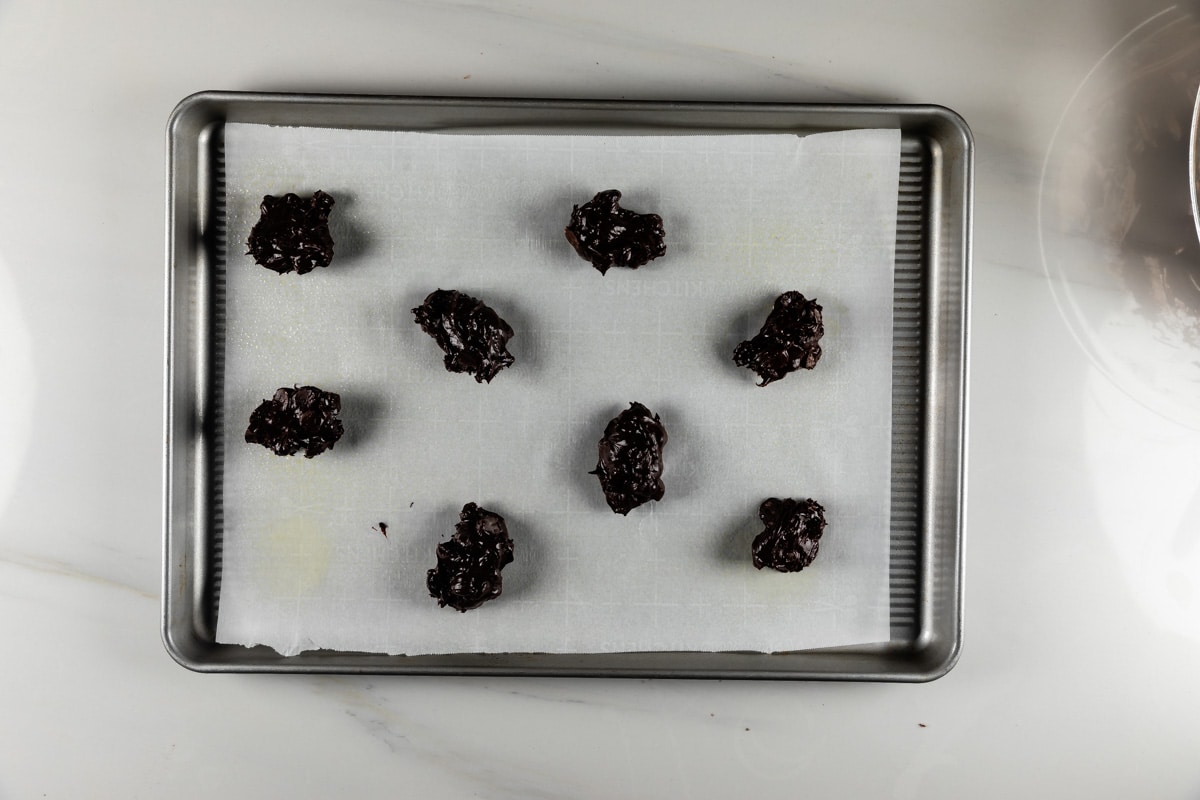 Eight dollops of dark brown cookie dough are arranged on a parchment-lined baking sheet, ready for the oven. The baking sheet rests on a light-colored countertop.