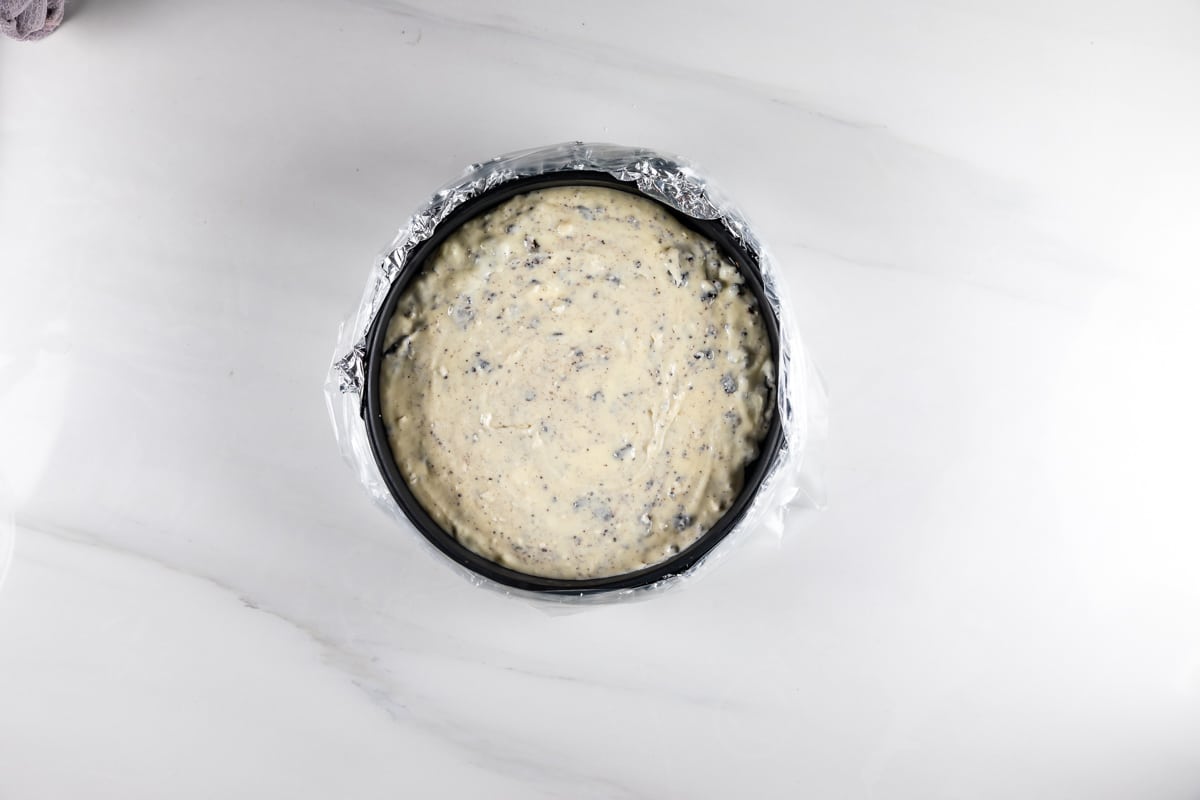 An overhead view of a cake pan lined with foil and filled with creamy cheesecake batter mixed with cookie pieces, set on a white marble surface.