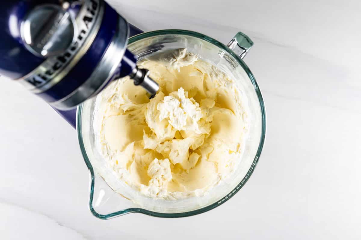 A stand mixer with a glass bowl is filled with whipped cream cheese and butter on a white countertop. The beaters are partially visible, indicating the blending process.