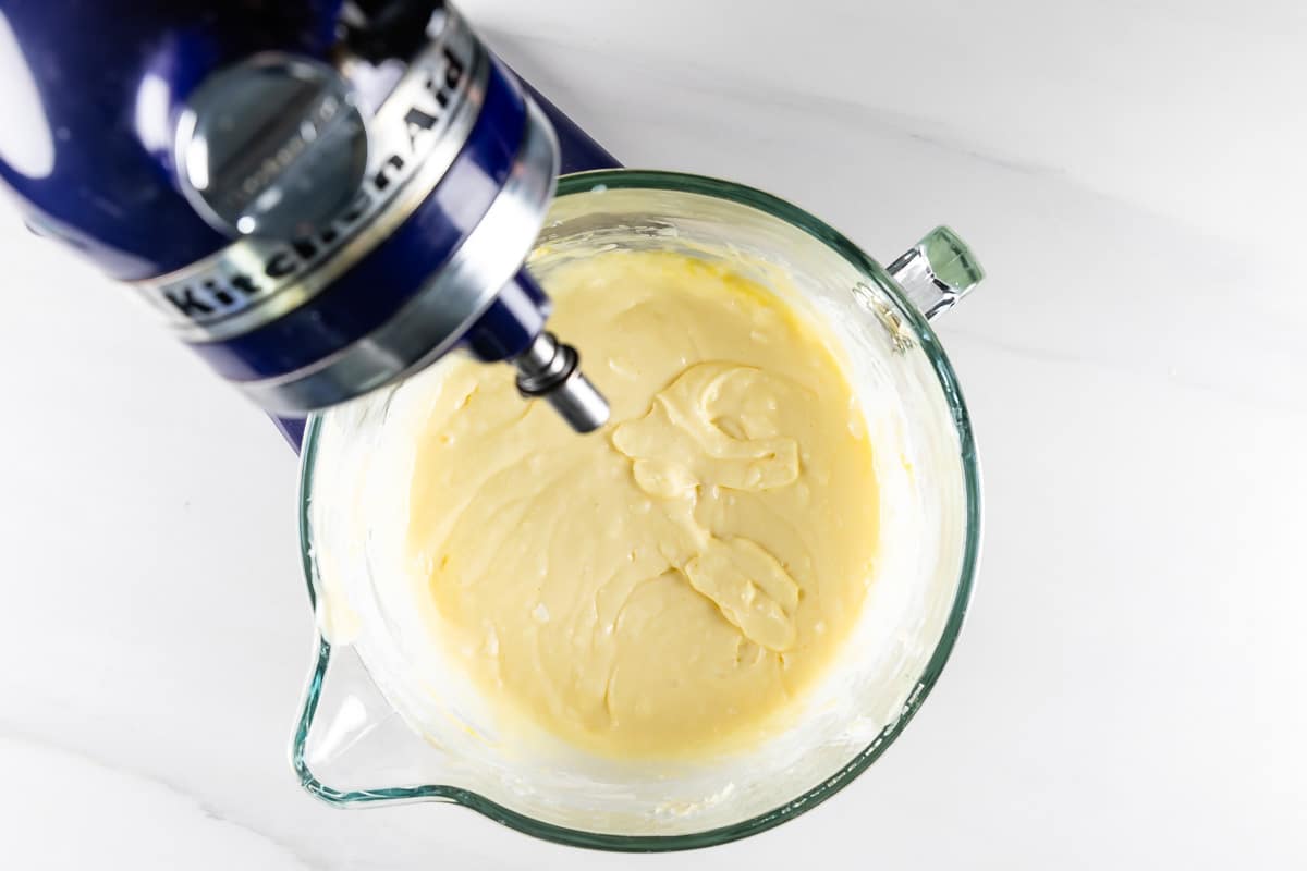 A top view of a stand mixer with a blue casing and a glass bowl filled with smooth, creamy cake batter. The mixer paddle is poised above the batter, suggesting the mixing process. The background is a white marble surface.