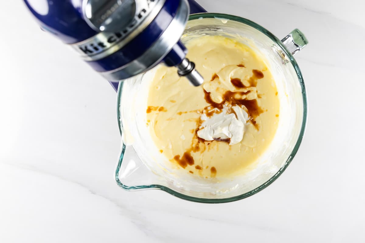 A glass mixing bowl sits beneath a blue stand mixer, containing creamy batter with swirls of cream cheese and vanilla. The bowl rests on a white countertop.