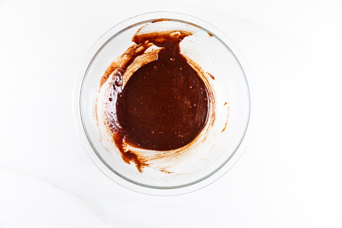 A glass bowl containing thick, dark brown chocolate batter with some streaks on the side, set on a white surface.