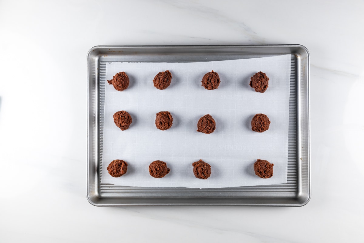 A baking tray with parchment paper holds twelve evenly spaced chocolate cookie dough balls, ready to be baked. The tray is placed on a white countertop.