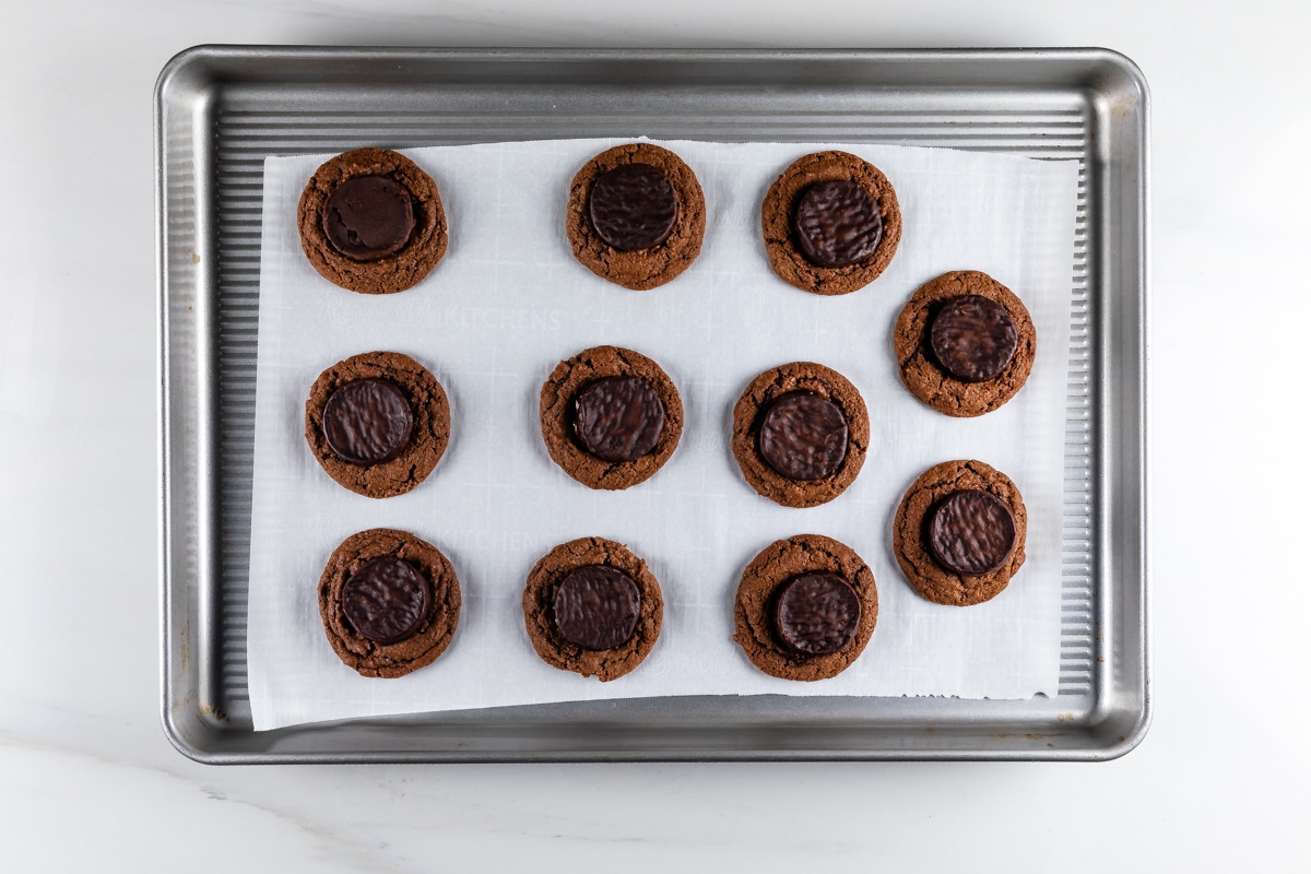 A baking tray holds ten chocolate thumbprint cookies with dark chocolate centers, arranged in rows on white parchment paper. The tray is placed on a white marble surface.