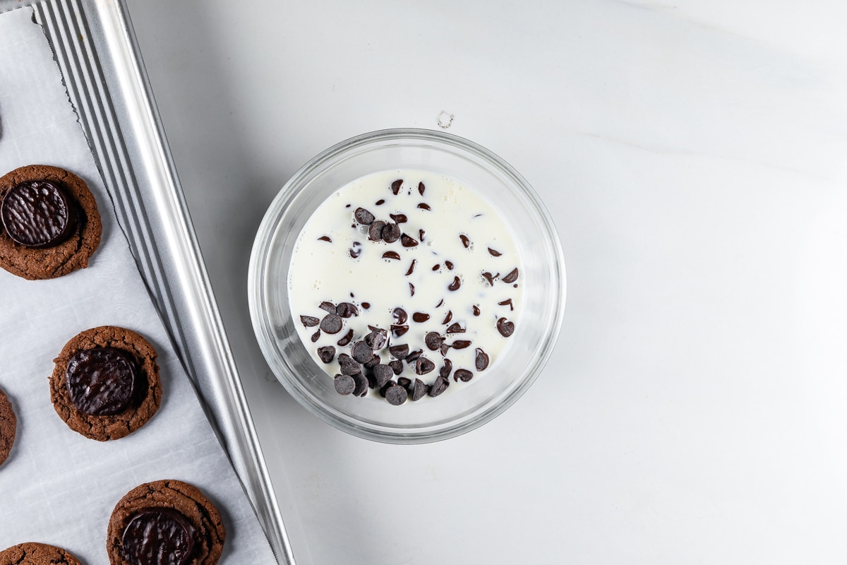 A bowl of milk with chocolate chips floating on top is placed on a white surface. To the left, a baking tray lined with parchment paper holds several chocolate cookies.