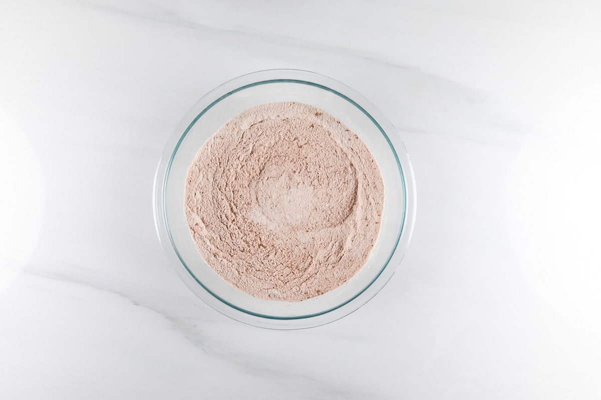 A glass bowl filled with beige powder sits on a white marble surface. The powder appears finely milled with a smooth, even texture.