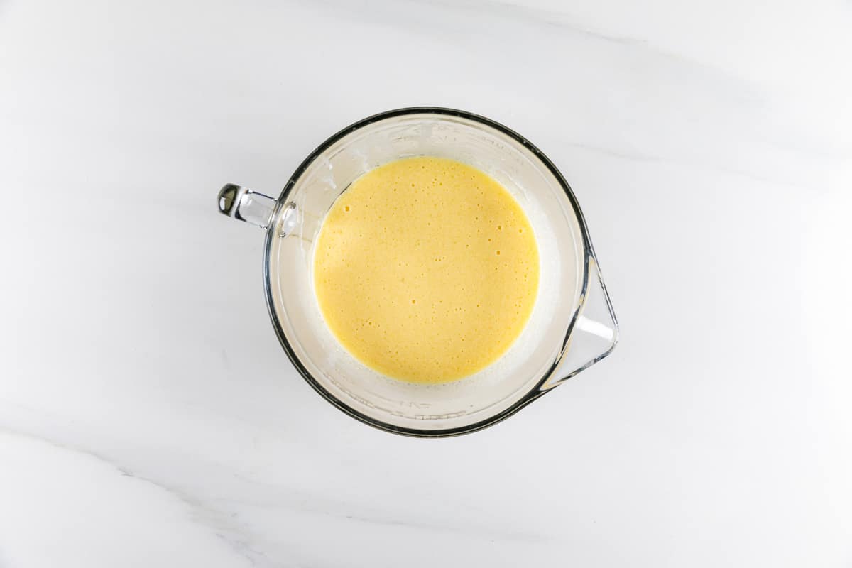 A glass bowl filled with a frothy, light yellow batter or mixture sits on a white marble surface. The image is taken from above, showing the smooth texture of the mixture.