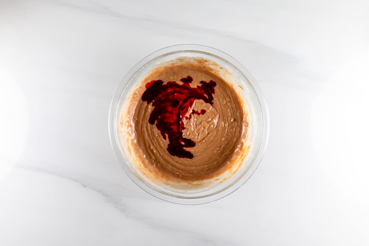 A glass bowl filled with light brown batter is placed on a white surface. Red liquid is drizzled on top of the batter, creating a contrast with the brown mixture. The image is bright and simple.