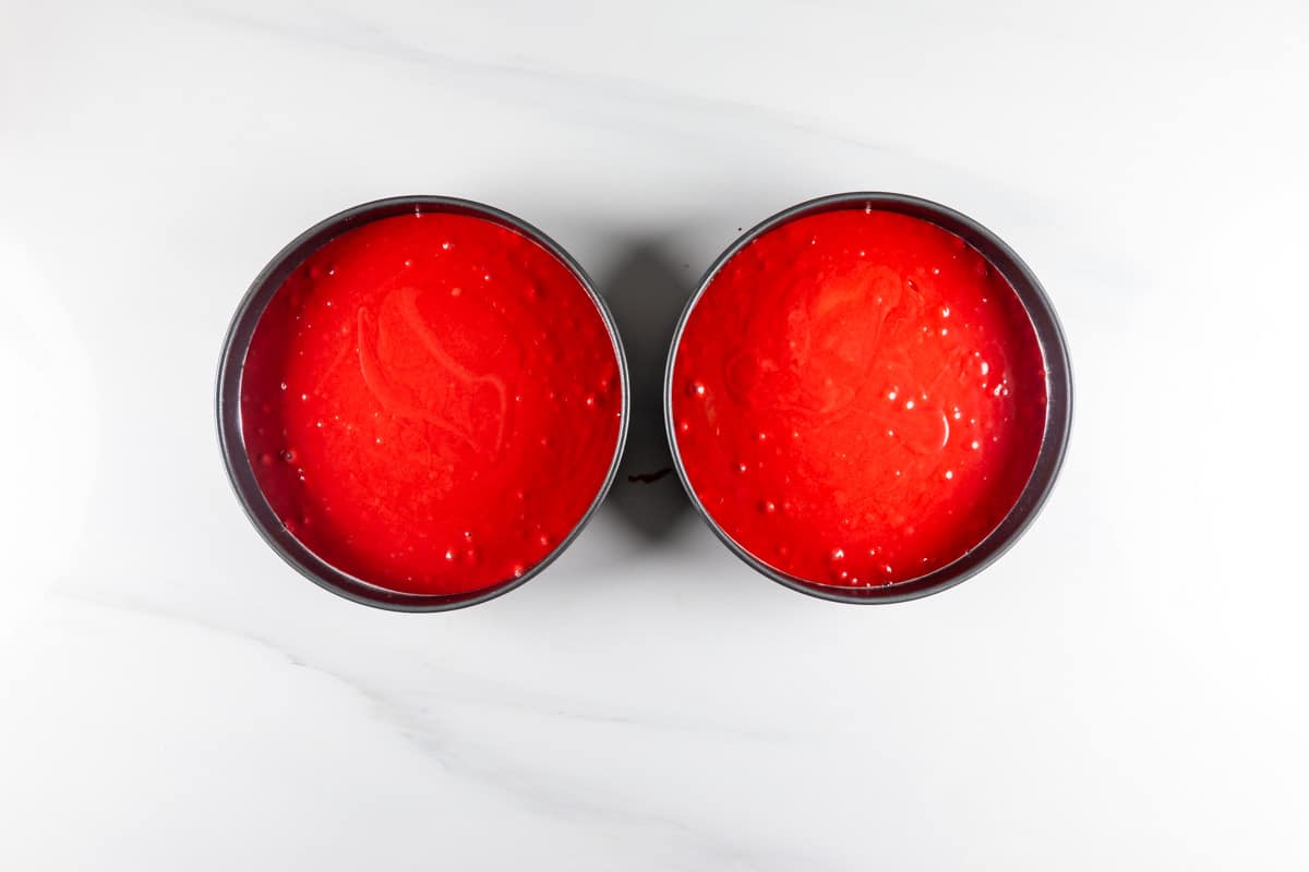 Two round baking pans filled with smooth, bright red cake batter are placed side by side on a white surface.