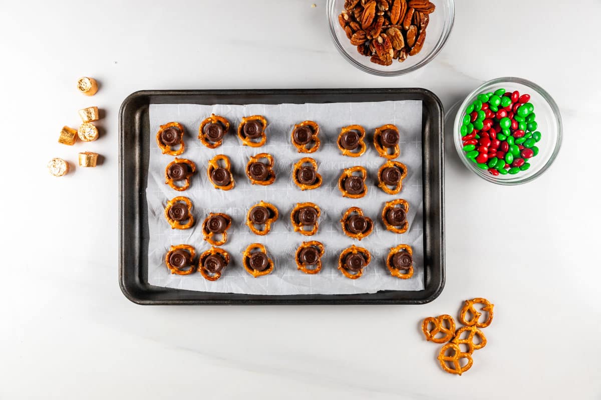 A baking tray with pretzel treats topped with chocolates and nuts, placed on a white surface. Surrounding the tray are bowls of pecans and red and green candies, along with scattered pretzels and chocolate pieces.