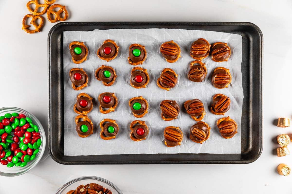 A baking tray filled with pretzels topped with melted candy and nuts, arranged in neat rows on parchment paper. Small bowls of red and green candies, pretzels, and pecans are placed nearby on the table.