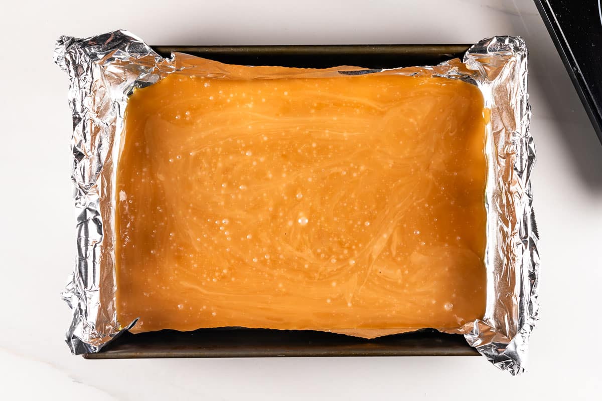 A baking tray lined with aluminum foil is filled with a smooth layer of caramel. The caramel has a glossy finish and is cooling on a white countertop.