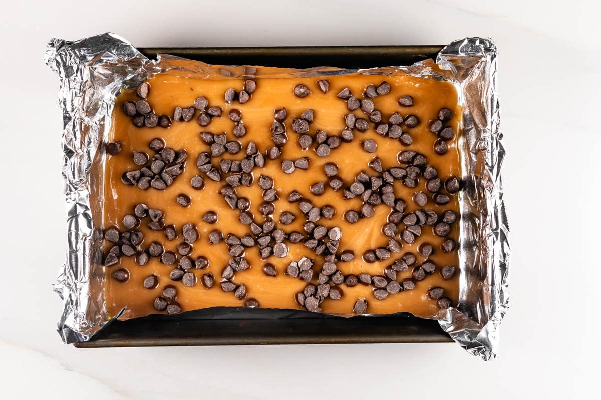 A rectangular baking pan lined with foil contains caramel-colored fudge topped with scattered chocolate chips. The dessert is set on a white surface.