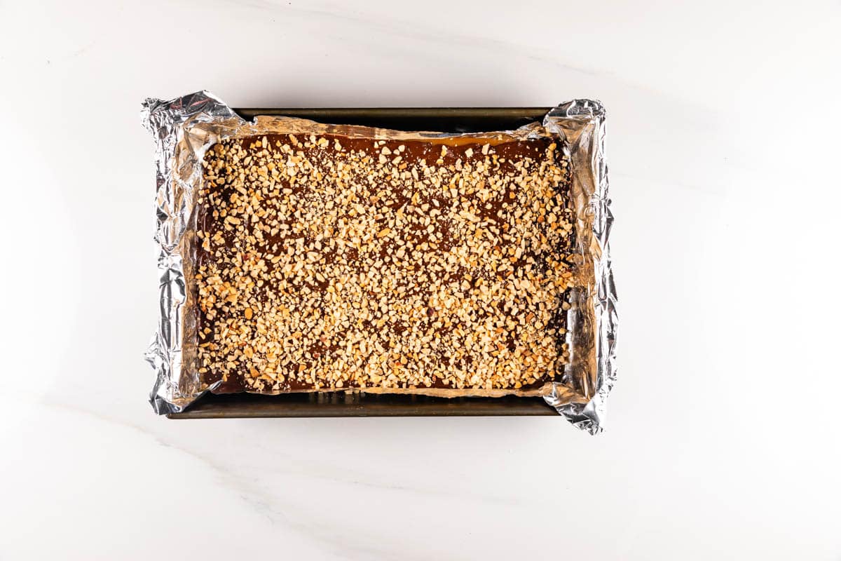 A rectangular baking tray lined with foil contains a dessert topped with a layer of crushed nuts. The dessert has a glossy finish and is evenly spread within the tray. The tray sits on a white surface.