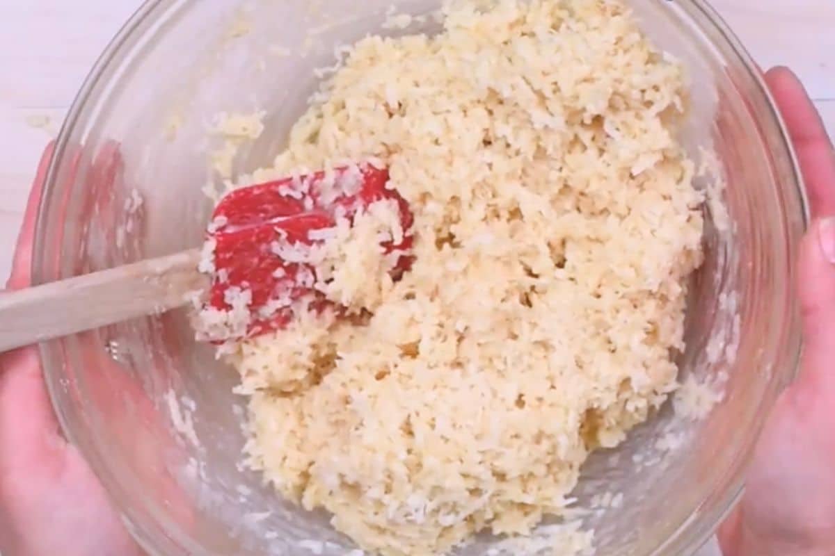 A person holds a glass bowl filled with a light, fluffy mixture. A red spatula is resting inside the bowl, partially covered by the mixture, which appears to be a batter or dough. The bowl is positioned over a light-colored surface.