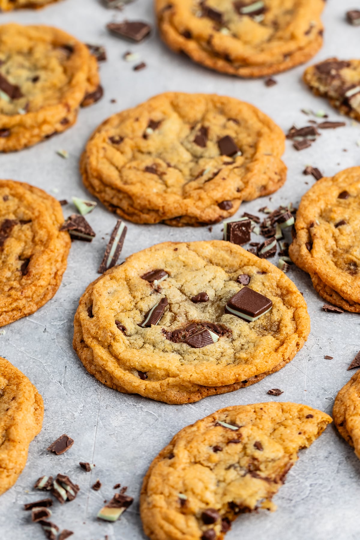 A batch of freshly baked chocolate chip cookies on a gray surface. Some cookies have chunks of chocolate scattered around them. One cookie has a bite taken out of it.