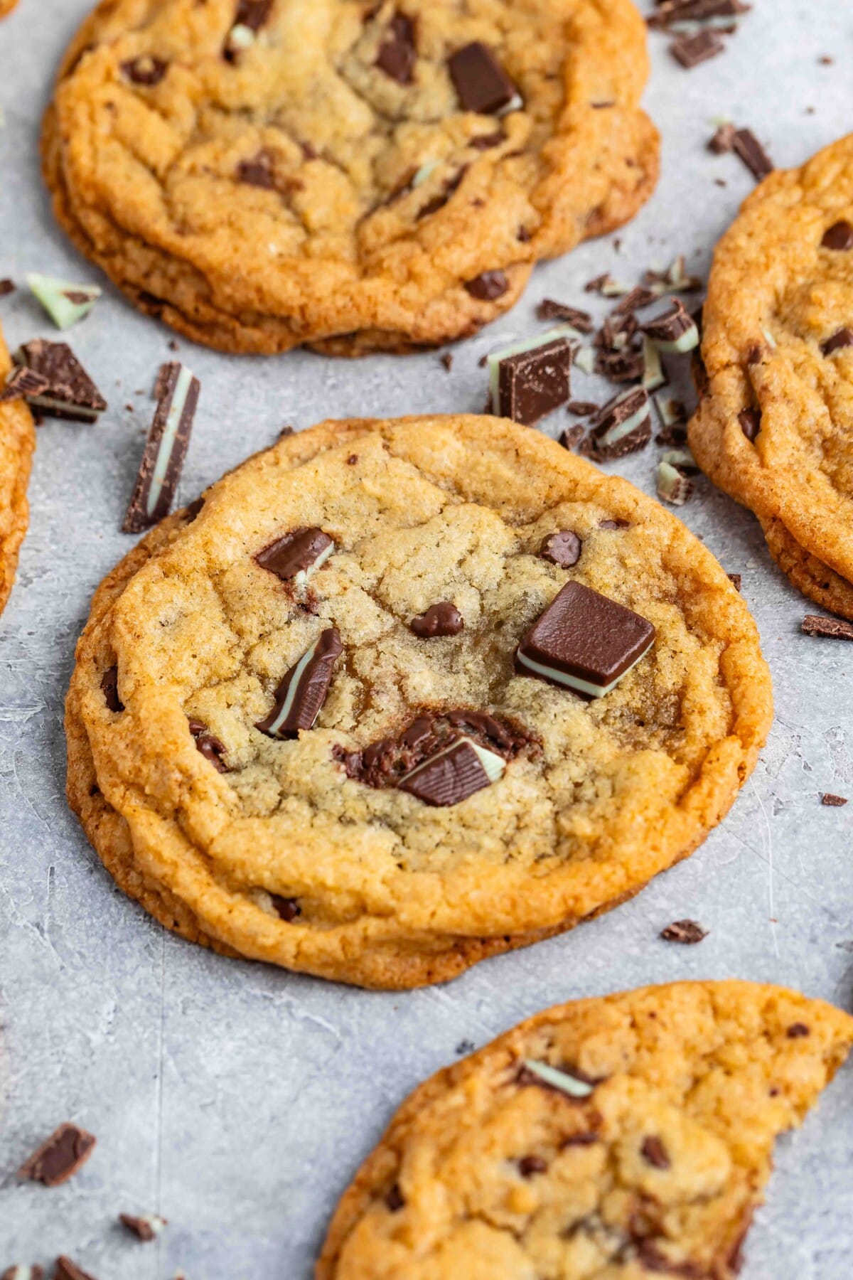 Close-up of several chocolate chip cookies on a light gray surface. The cookies are golden-brown, with chunks of chocolate on top and around them, suggesting freshness and a soft texture.