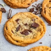 Close-up of several chocolate chip cookies on a light gray surface. The cookies are golden-brown, with chunks of chocolate on top and around them, suggesting freshness and a soft texture.