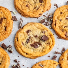 A group of freshly baked chocolate chip cookies with chunks of chocolate on top, arranged on a light gray surface. Some chocolate pieces are scattered around, enhancing the homemade and cozy appearance of the cookies.