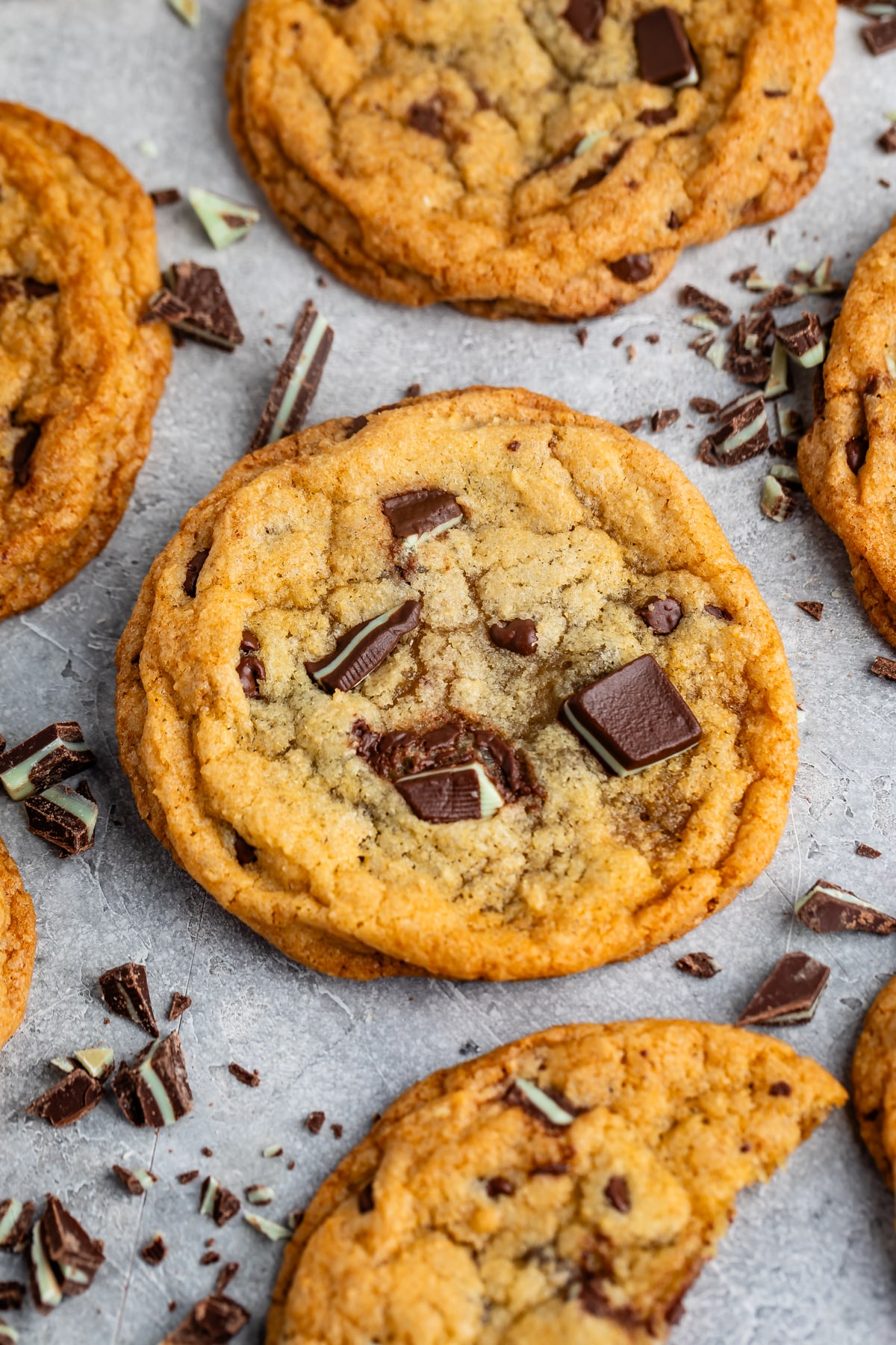 Close-up of chocolate chip cookies on a gray surface. The cookies are golden brown, with chunks of chocolate scattered around them. Some chocolate pieces are embedded in the cookies, giving a freshly baked appearance.