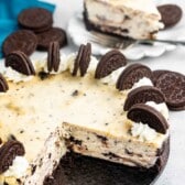 A sliced Oreo cheesecake on a black plate, topped with whipped cream and Oreo halves. A piece is served on a white plate in the background, next to a stack of whole Oreo cookies. A teal napkin is nearby.