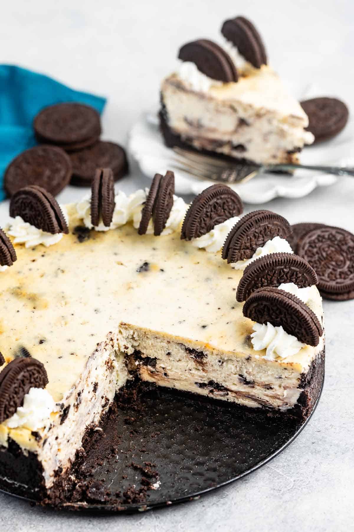 A sliced Oreo cheesecake on a black plate, topped with whipped cream and Oreo halves. A piece is served on a white plate in the background, next to a stack of whole Oreo cookies. A teal napkin is nearby.