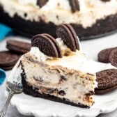 A slice of Oreo cheesecake on a white plate with whipped cream and Oreo cookies on top. The cheesecake has a chocolate cookie crust and visible chocolate chunks. More whole Oreo cookies are scattered around the plate. A fork is beside the slice.