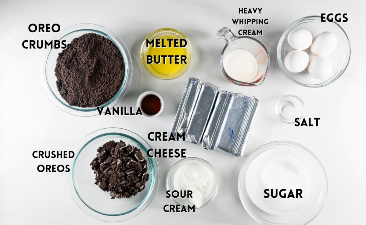 An overhead view of cheesecake ingredients on a white surface: Oreo crumbs, crushed Oreos, vanilla, melted butter, cream cheese, sour cream, heavy whipping cream, eggs, sugar, and salt, each labeled in separate containers.