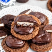 A plate of chocolate-covered cookies is stacked, with one cookie in the center broken in half to reveal a white filling. The cookies have a glossy chocolate coating and sit on a decorative white plate. Some wrapped cookies are blurred in the background.