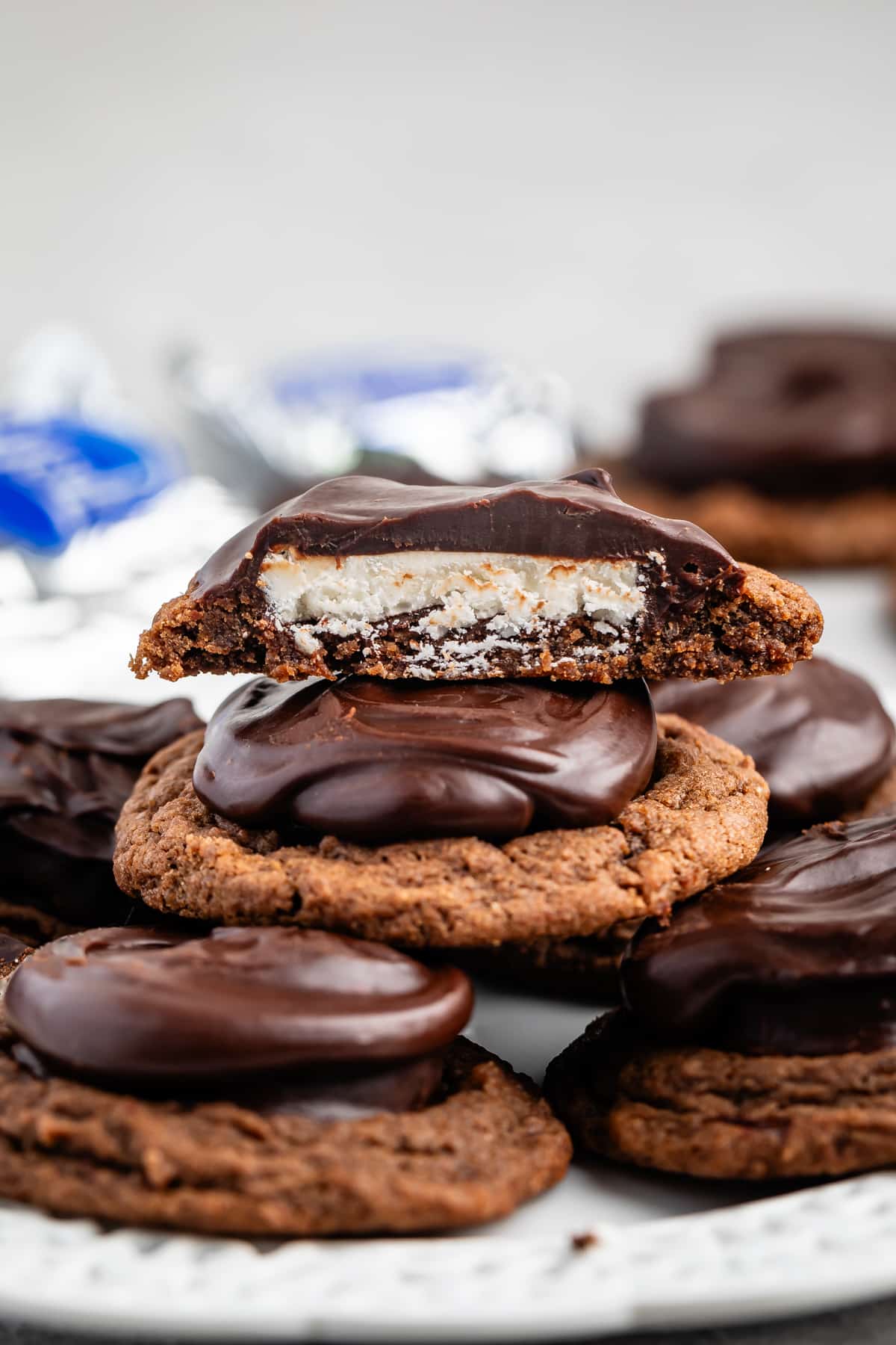 A stack of chocolate-covered cookies, with the top cookie bitten to reveal a cream filling. Several whole cookies are arranged around the stack on a white plate. Silver-wrapped candies are blurred in the background.