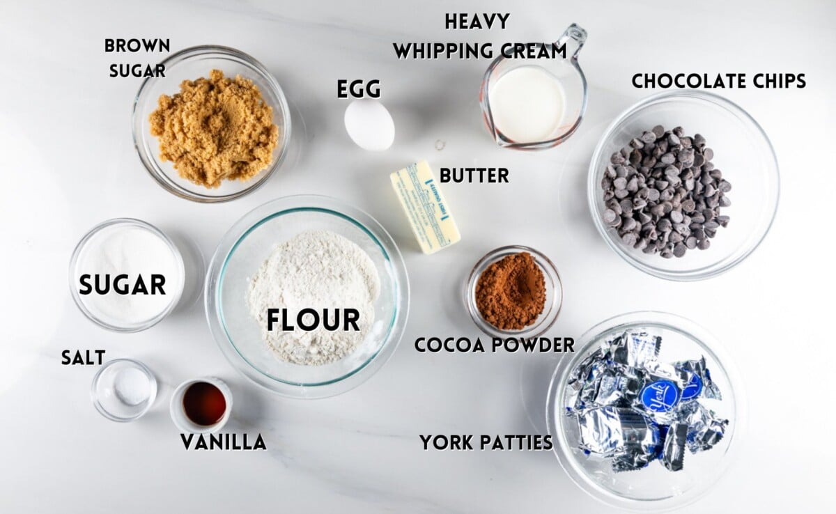 Top-down view of dessert ingredients on a white surface, labeled: brown sugar, sugar, flour, salt, vanilla, egg, butter, heavy whipping cream, cocoa powder, chocolate chips, and York patties.